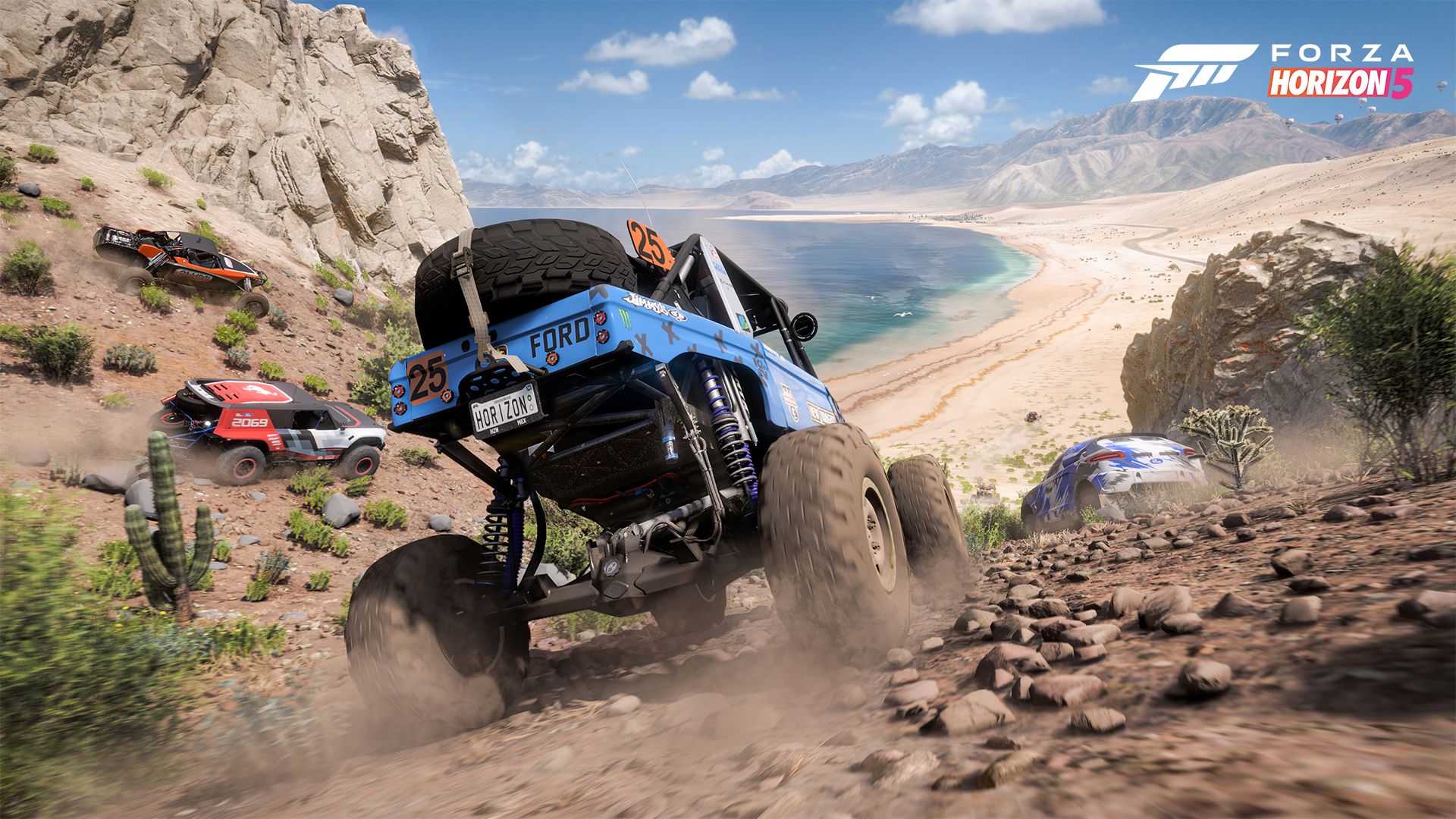 In the landscape of the Horizon Mexico Festival, four off-road vehicles speed down rocky desert hills toward a sandy beach on a clear, sunny day.