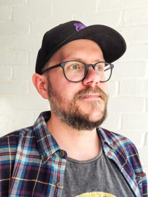 Profile image of artist Steve Wade wearing a baseball hat, black-rimmed glasses and a plaid shirt.