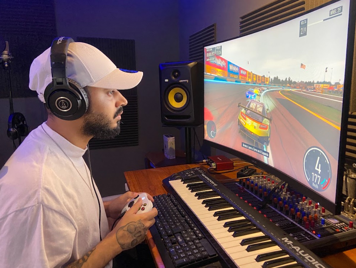 Wzy, um homem vestindo um bonê branco, camiseta branca e com fones, está sentado em frente a um monitor, segurando um controle de Xbox enquanto joga Forza